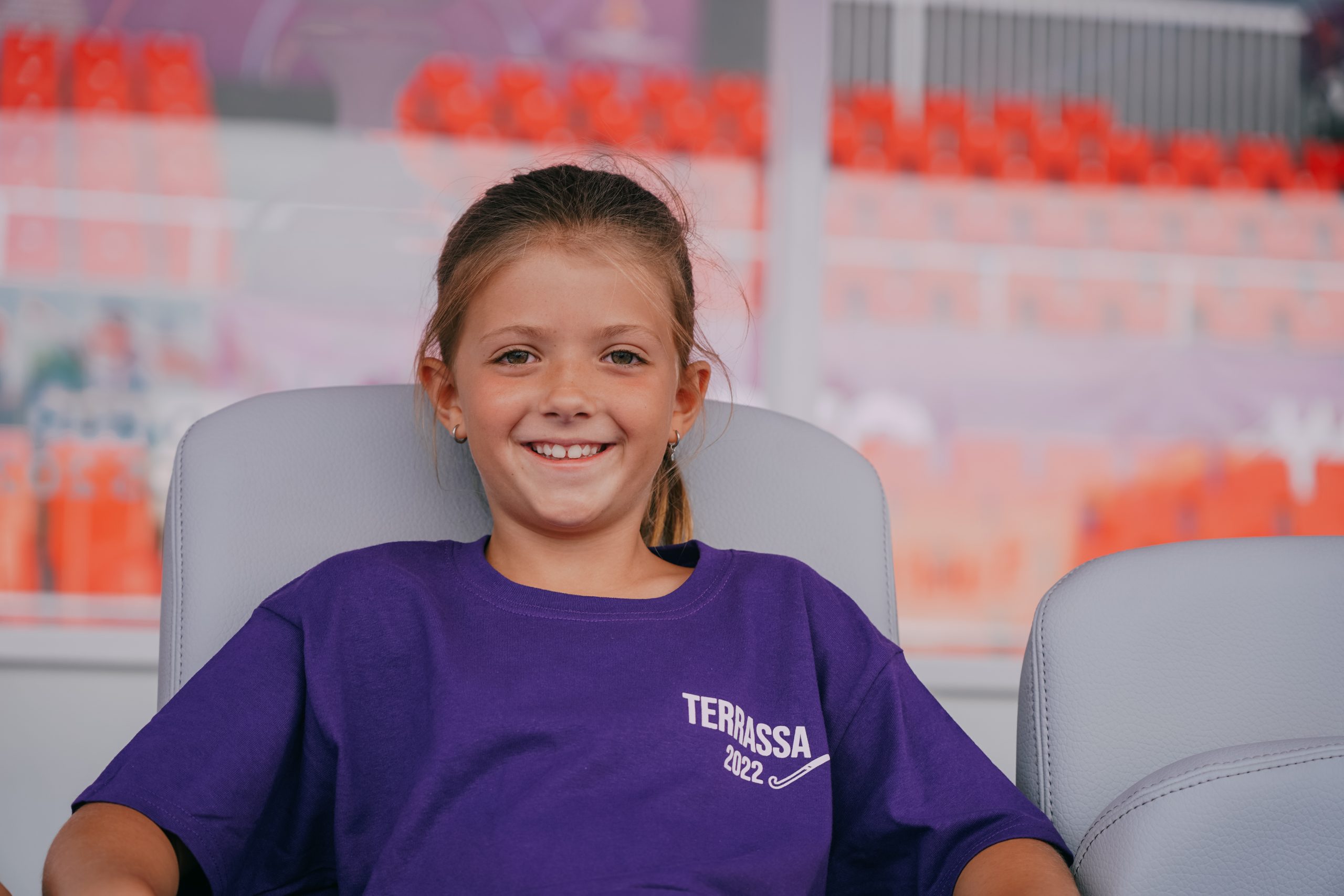 niña sonriendo sentada en el banquillo de un campo de hockey con una camiseta unisex Kukuxumusu de algodón de color morado con el dibujo de un palo de hockey y la palabra TERRASSA 2022 en el pecho